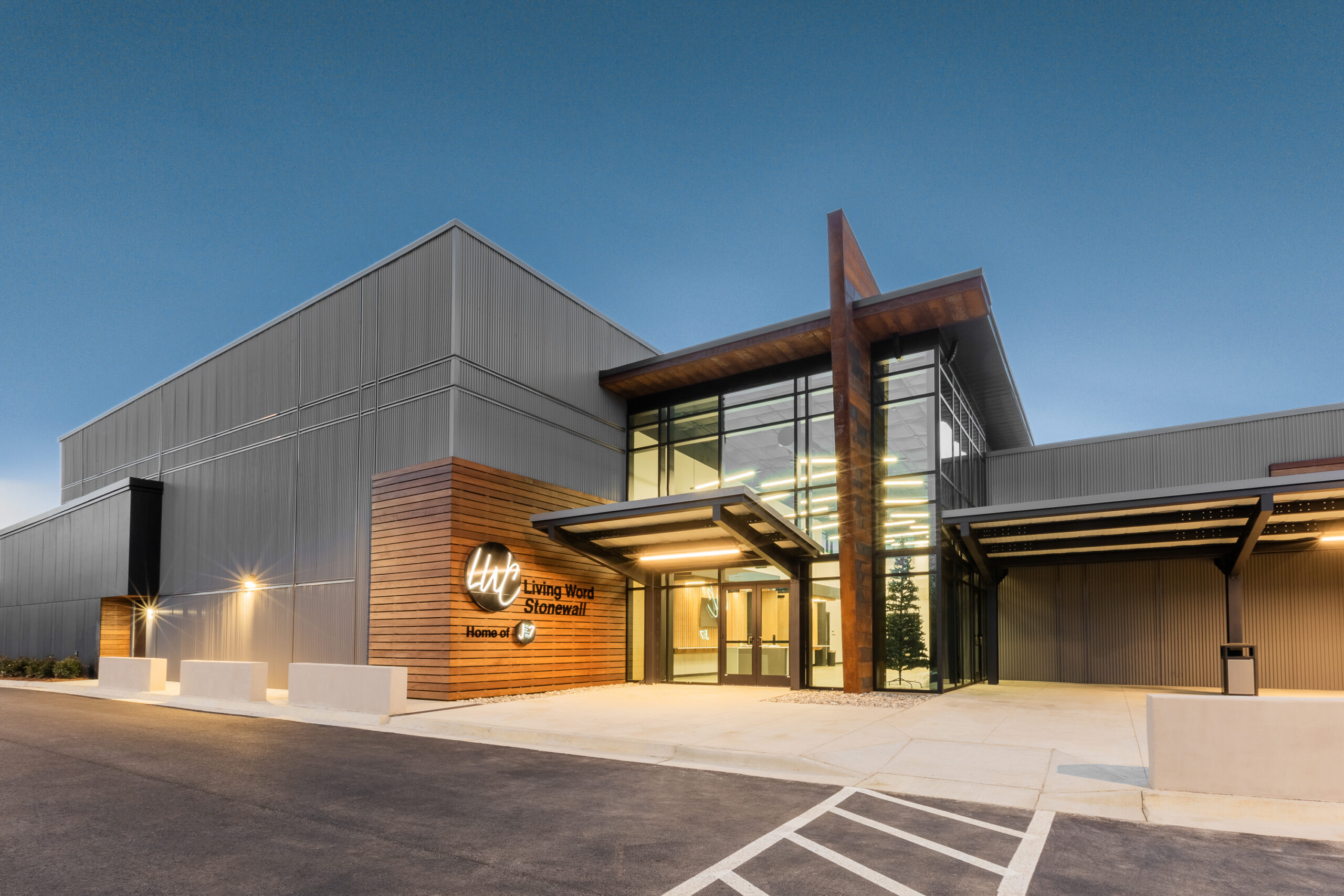 Twilight exterior of Living Word Church Stonewall with modern dark metal cladding wood accents cross and illuminated entry