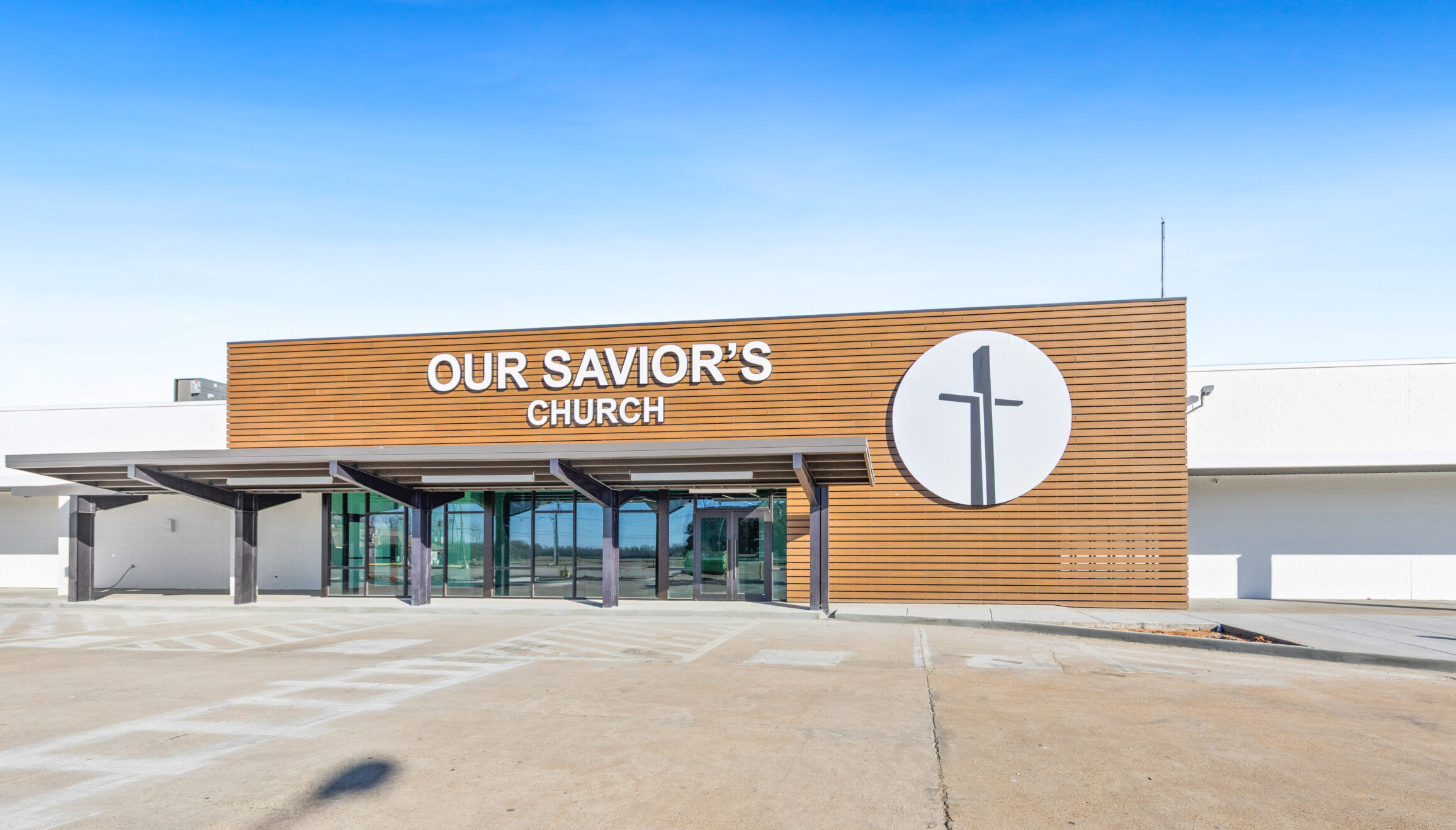 Modern exterior of Our Saviour's Church Ville Platte campus featuring contemporary composite wood slat siding, illuminated cross logo, and cantilevered entry canopy designed by A Beazley Architecture